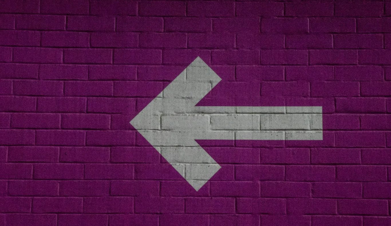 Left arrow symbol against a purple brick wall background.